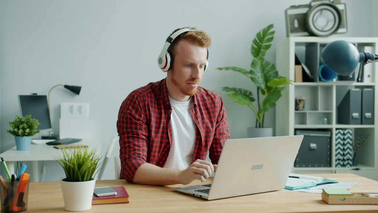 A person wearing headphones is focused on a laptop in a modern home office. The desk has plants and books, with shelves in the background, conveying a productive mood.