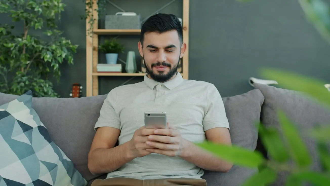 A man with a beard sits on a gray sofa and smiles while looking at his silver smartphone in a modern living room setting.