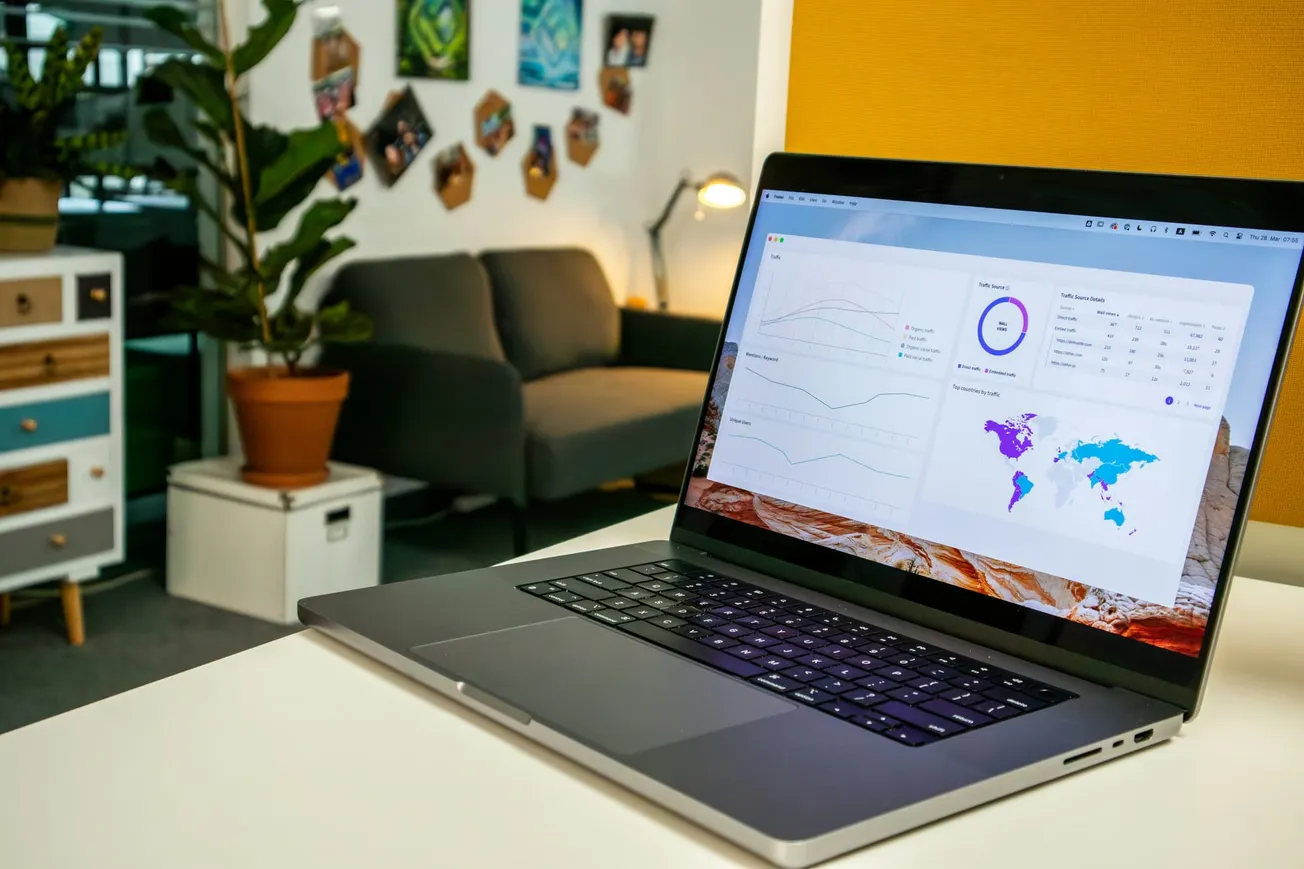 Laptop displaying analytics graphs and maps on a desk in a cozy office with a gray sofa, potted plant, colorful drawer, and photos on the wall.
