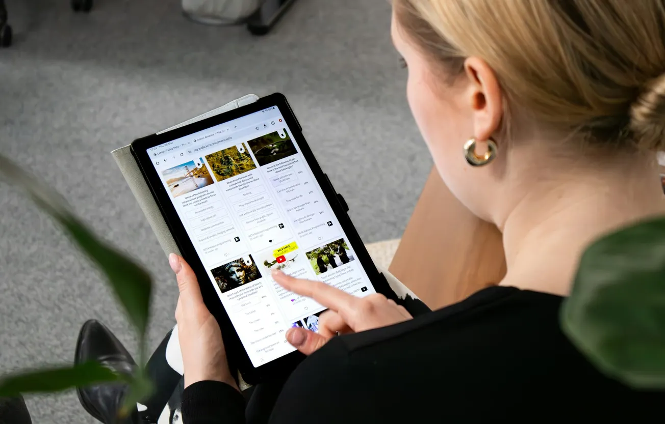 A woman with earrings uses a tablet, browsing travel destinations with images and text. Her focus suggests curiosity and engagement. Background shows a carpet floor.