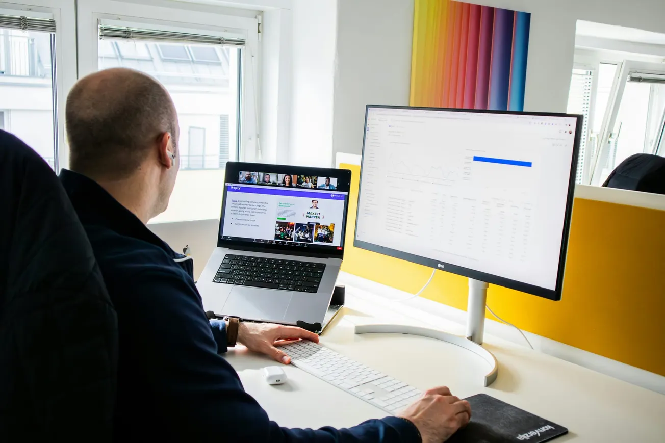 A person in an office setting works at a desk with a laptop and a large monitor displaying graphs. There’s a colorful artwork on the wall, giving a focused yet creative atmosphere.