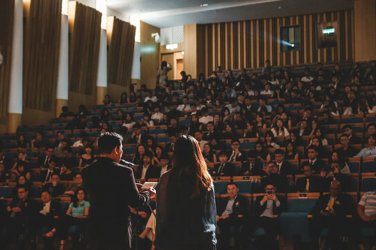 Two people address a large, seated audience in a dimly lit auditorium, creating an atmosphere of focused attention and engagement.