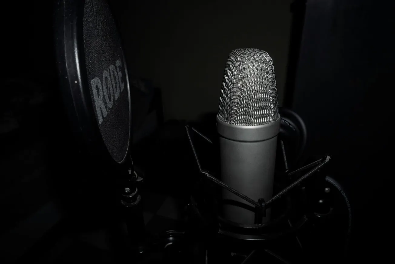 Close-up of a studio microphone with a pop filter, set against a dark background. The mic is silver with a textured grille.