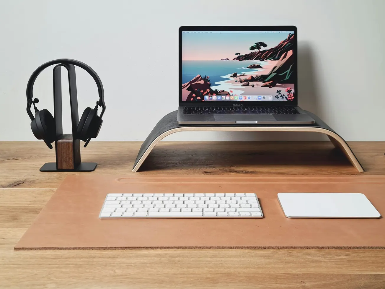 Minimalist workspace featuring a laptop on a wooden stand, a sleek pair of headphones on a stand, a keyboard, and trackpad on a wooden desk.