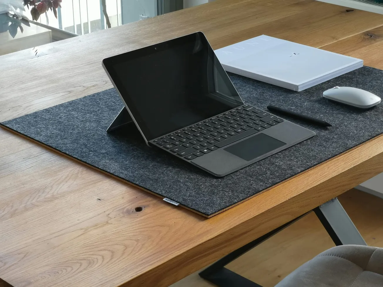 A sleek tablet with a keyboard and stylus rests on a black desk mat on a wooden table, accompanied by a closed laptop and mouse, evoking a modern workspace.