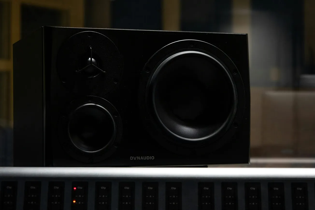 A black Dynaudio speaker with large circular elements sits on a sound mixing console, conveying a professional audio studio vibe.