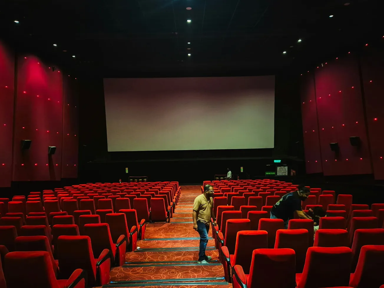 Empty cinema with red seats and large blank screen. Dim lighting sets a quiet, anticipatory mood. Two people are cleaning the aisles.