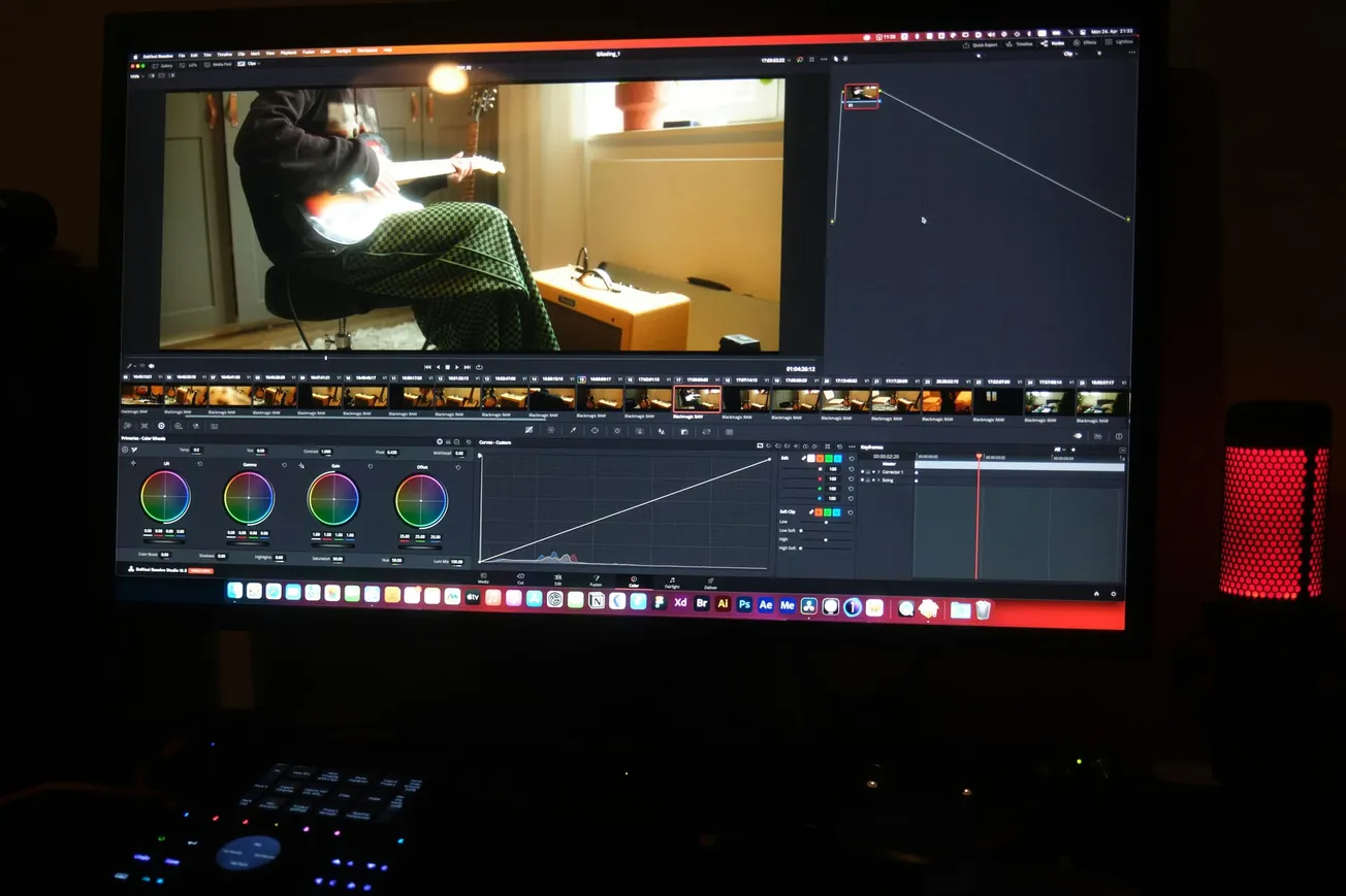 A computer monitor displays video editing software with color grading wheels, timeline, and video preview. A person plays guitar onscreen. A red microphone is nearby.