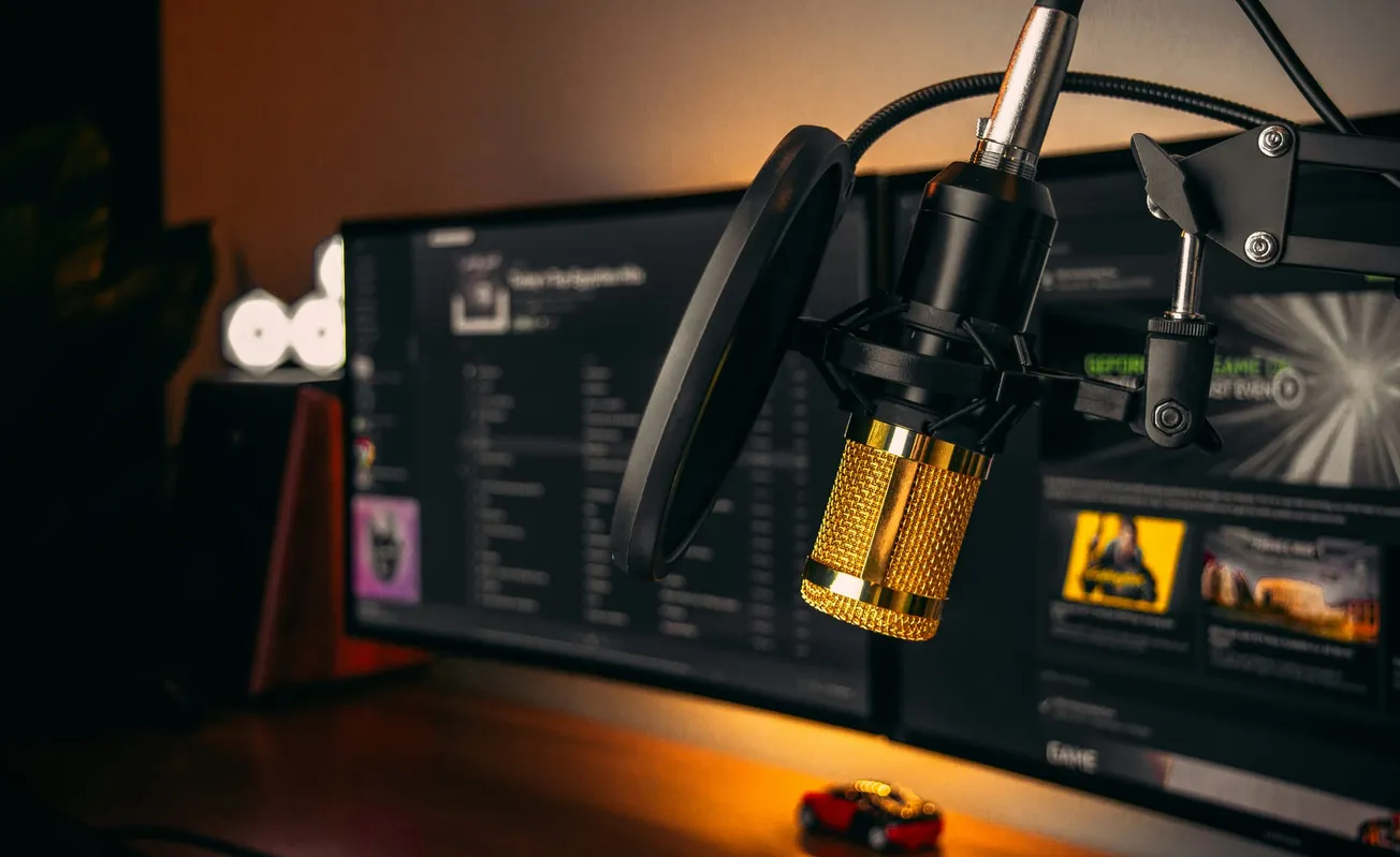 Close-up of a golden microphone and pop filter on an adjustable stand. Behind, blurred computer screens display music tracks, creating a cozy, tech-savvy atmosphere.