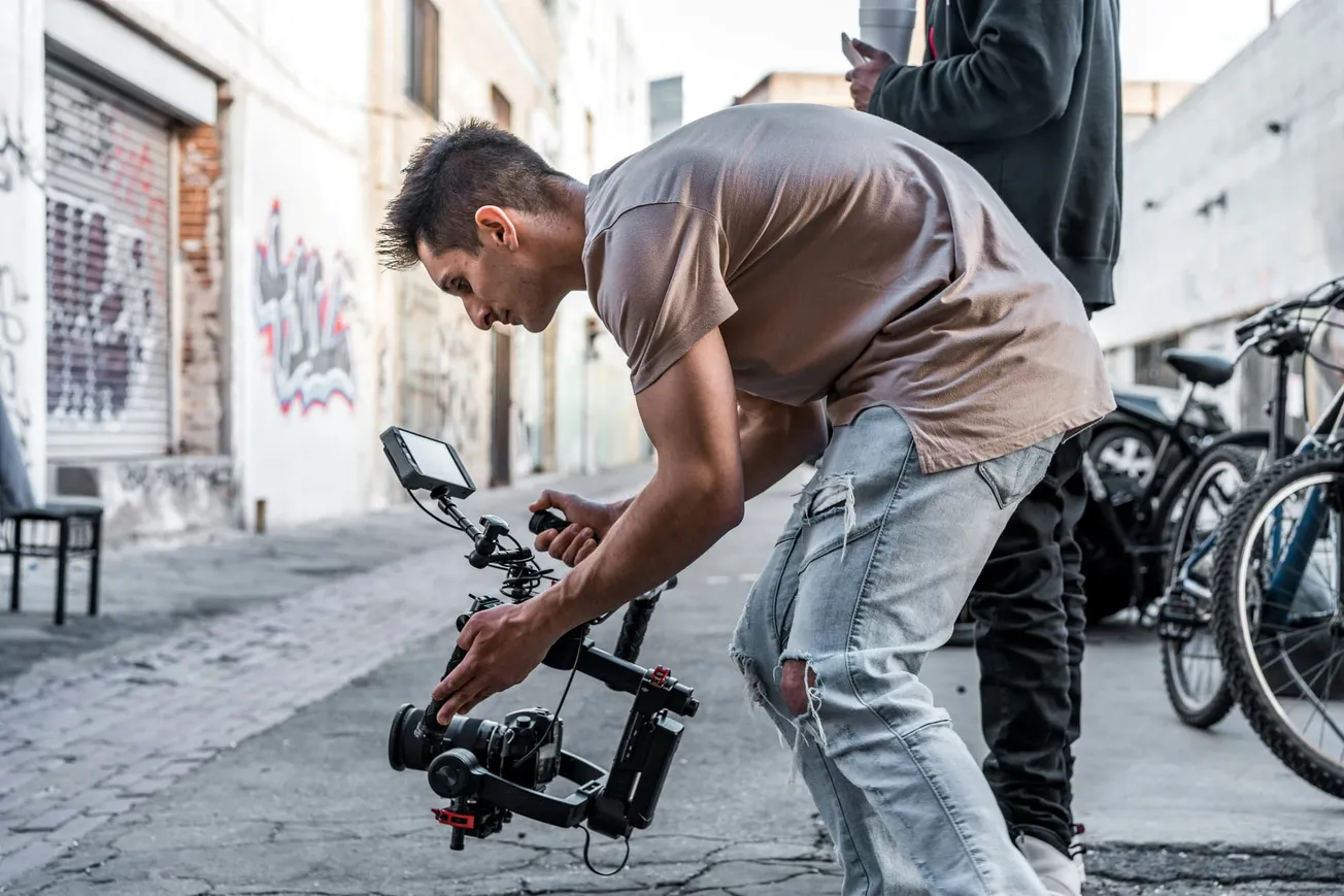 A man leans forward, operating a camera gimbal on a graffiti-lined urban street. He wears a casual t-shirt and jeans, concentrating intently.