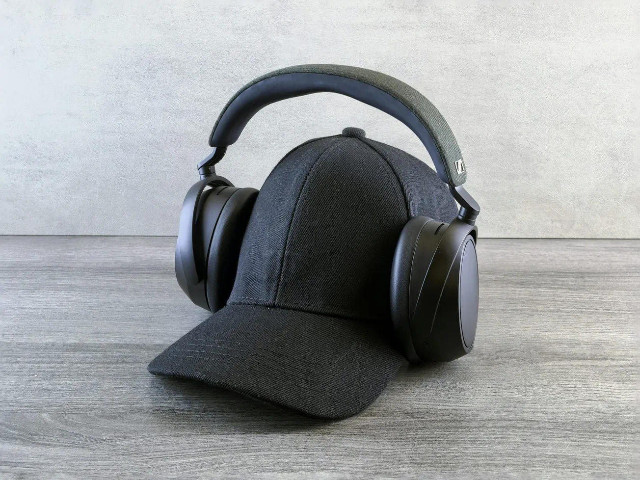 A black baseball cap is positioned on a wooden surface, with large black over-ear headphones resting on it, creating a modern, minimalist vibe.