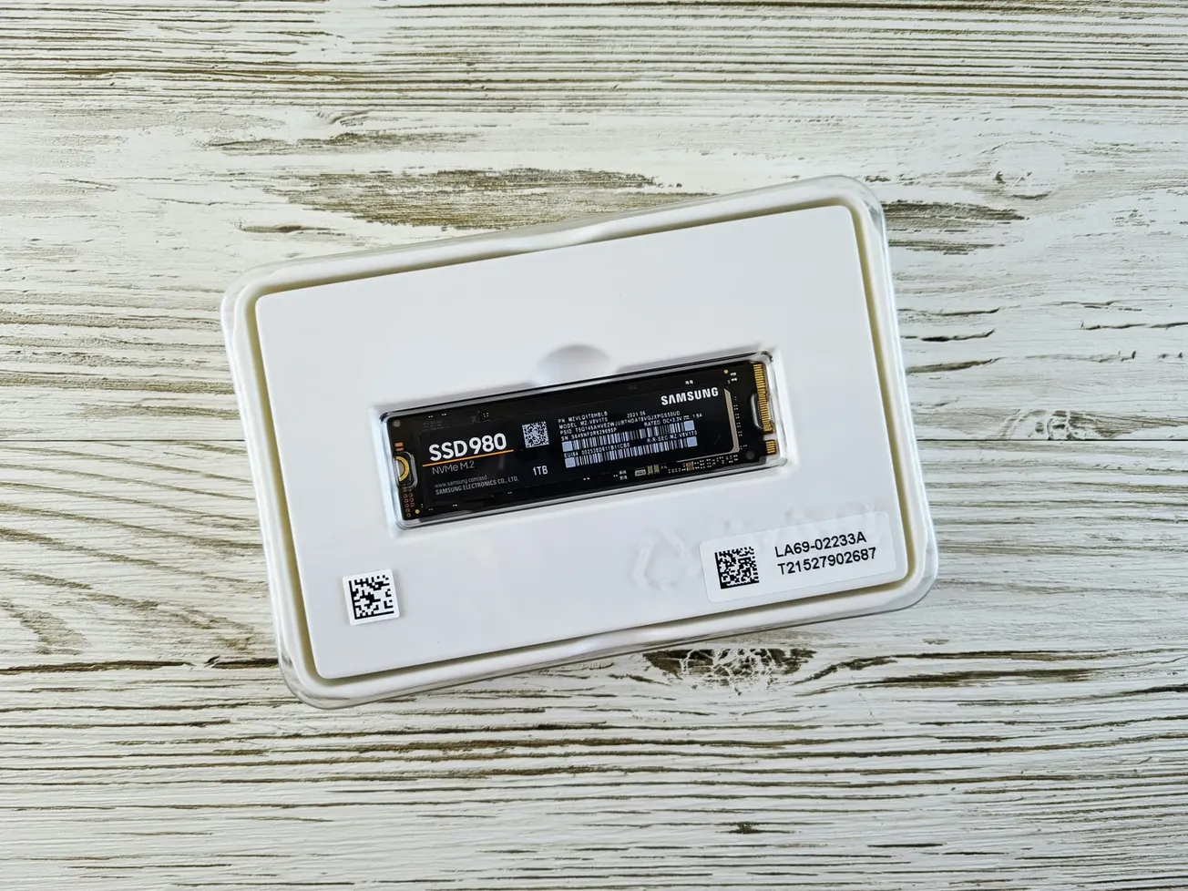 A Samsung SSD 980 lies on its white packaging, atop a textured wooden surface. The black drive features text and QR codes, giving a modern tech feel.
