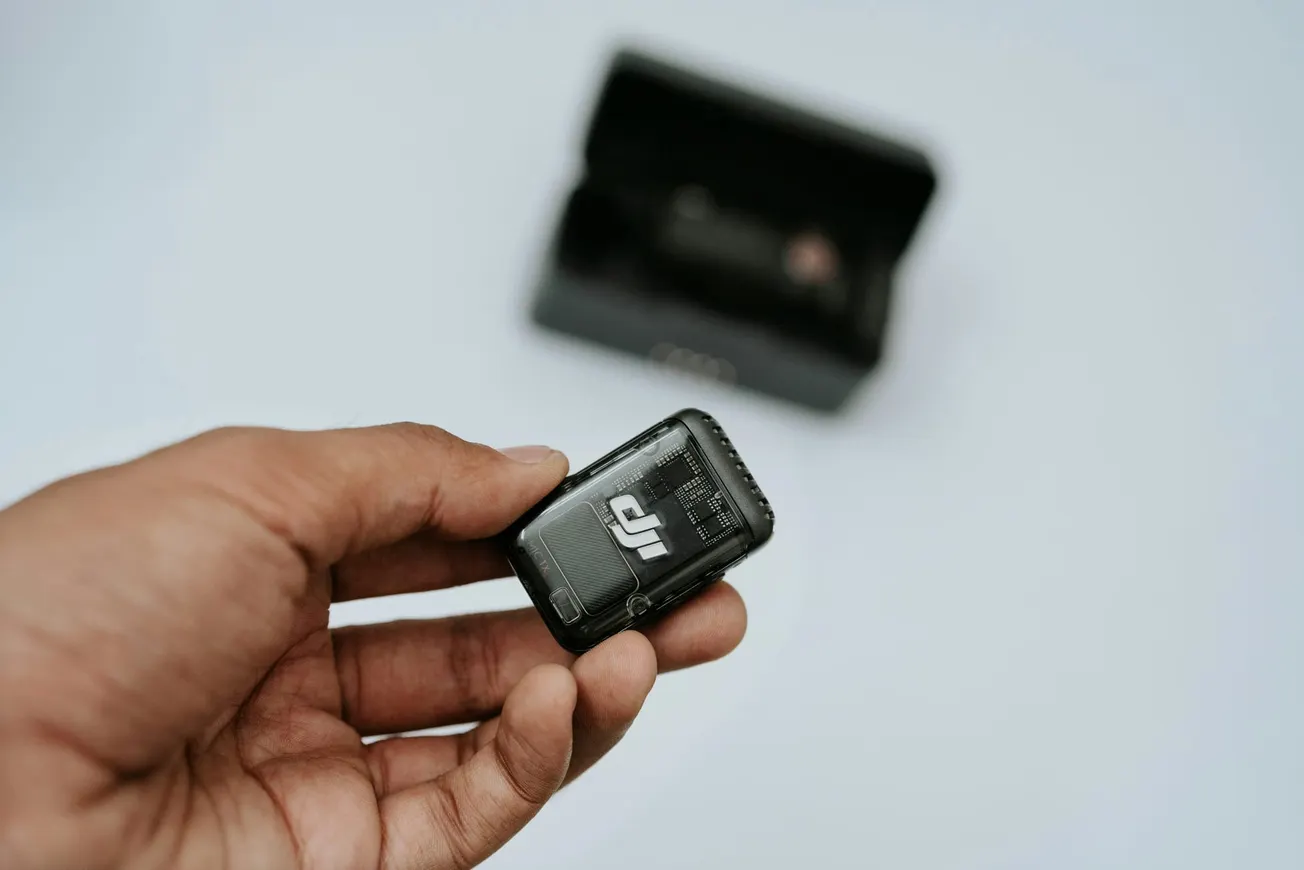A hand holding a small black electronic device with a logo, against a blurred background of an open black box. The tone is minimalist and tech-focused.