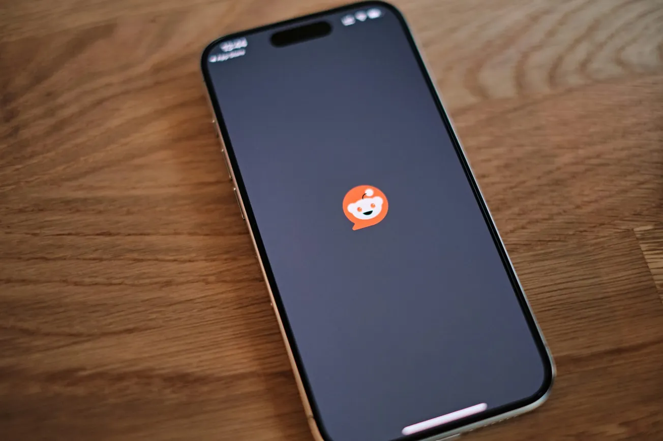 Smartphone on a wooden table displaying a loading screen with Reddit's orange logo centered on a black background, indicating the app is opening.