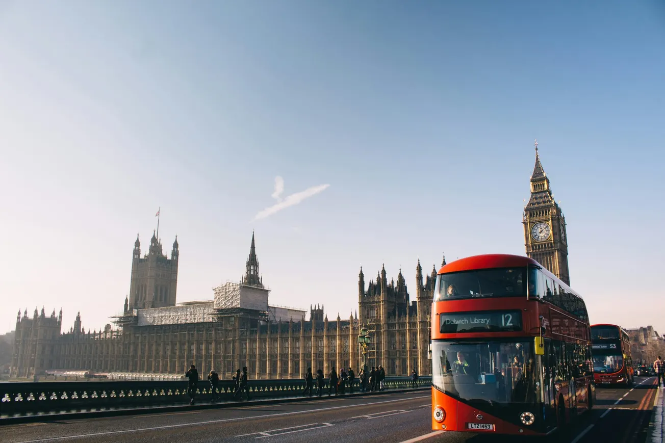 Red double-decker buses drive past the Houses of Parliament and Big Ben in London under a clear blue sky, capturing a lively city vibe.