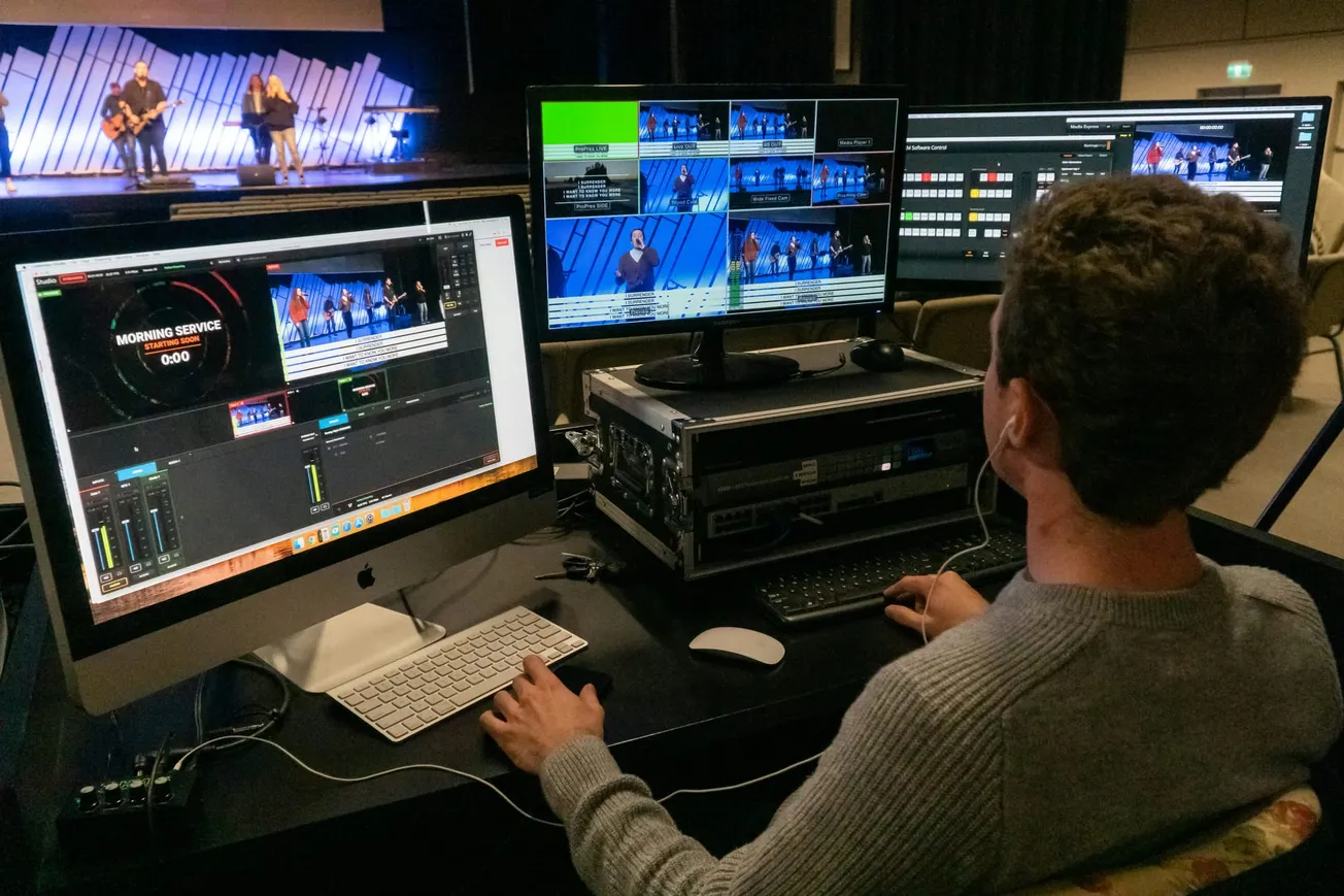 A person with curly hair operates a live-streaming setup, monitoring multiple screens showing a stage with musicians. The setting conveys a focused, technical atmosphere.