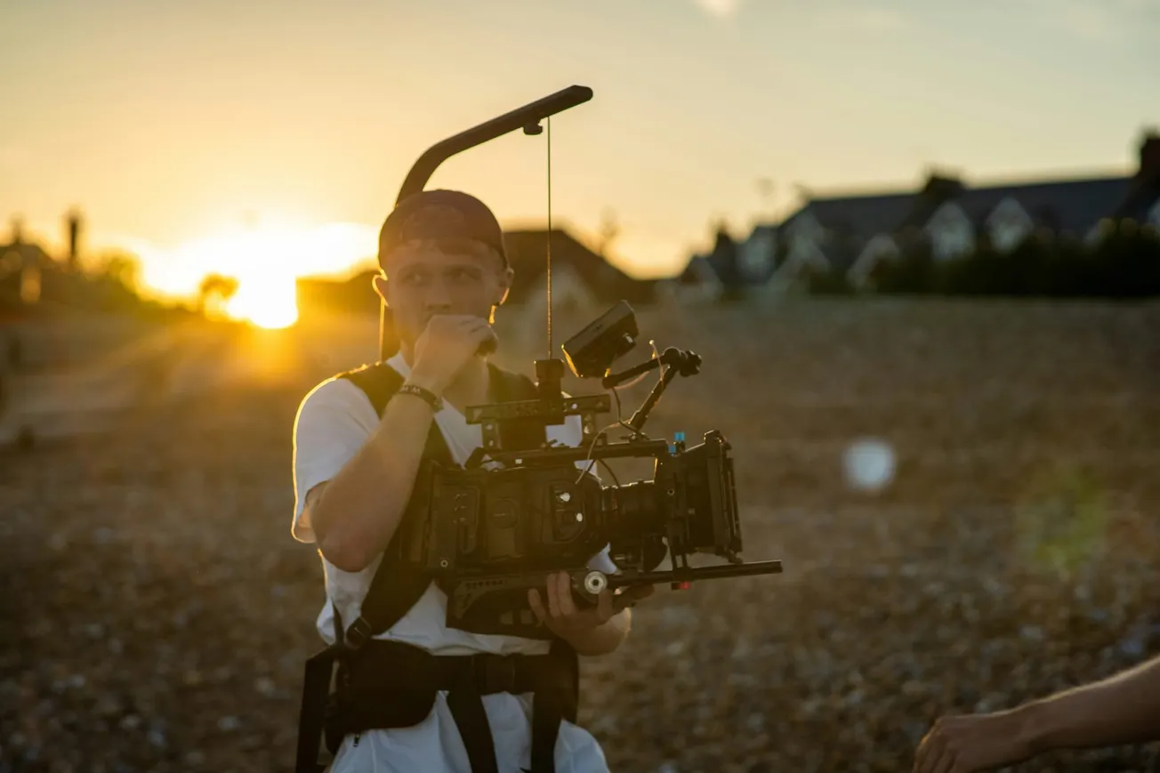 Cameraperson filming a scene at sunset on a pebble beach, with sun glowing behind them. They wear a harness, focused and absorbed in their work.