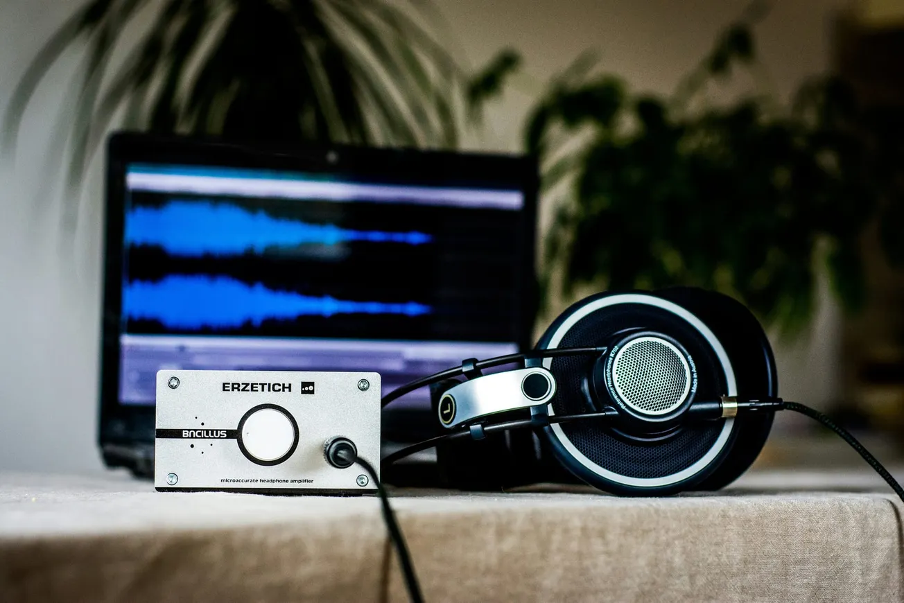Headphones rest on a table beside an audio amplifier, with a laptop showing sound waves in the background. The scene conveys a focus on music production.