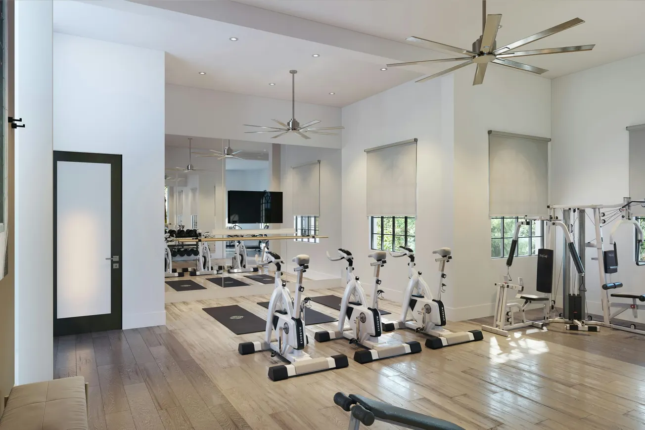 Modern gym with wooden floors, white walls, and large mirrors. There are stationary bikes, weight machines, and yoga mats under ceiling fans. Bright and spacious.