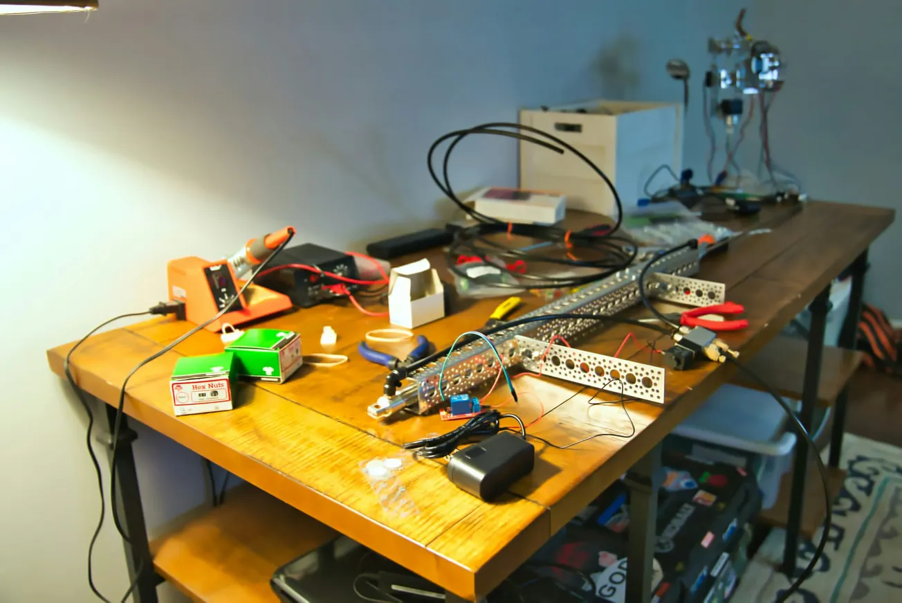A cluttered workbench with electronic components, wires, and tools. A soldering iron is to the left, with scattered boxes and cables. The scene conveys a busy, creative workspace.