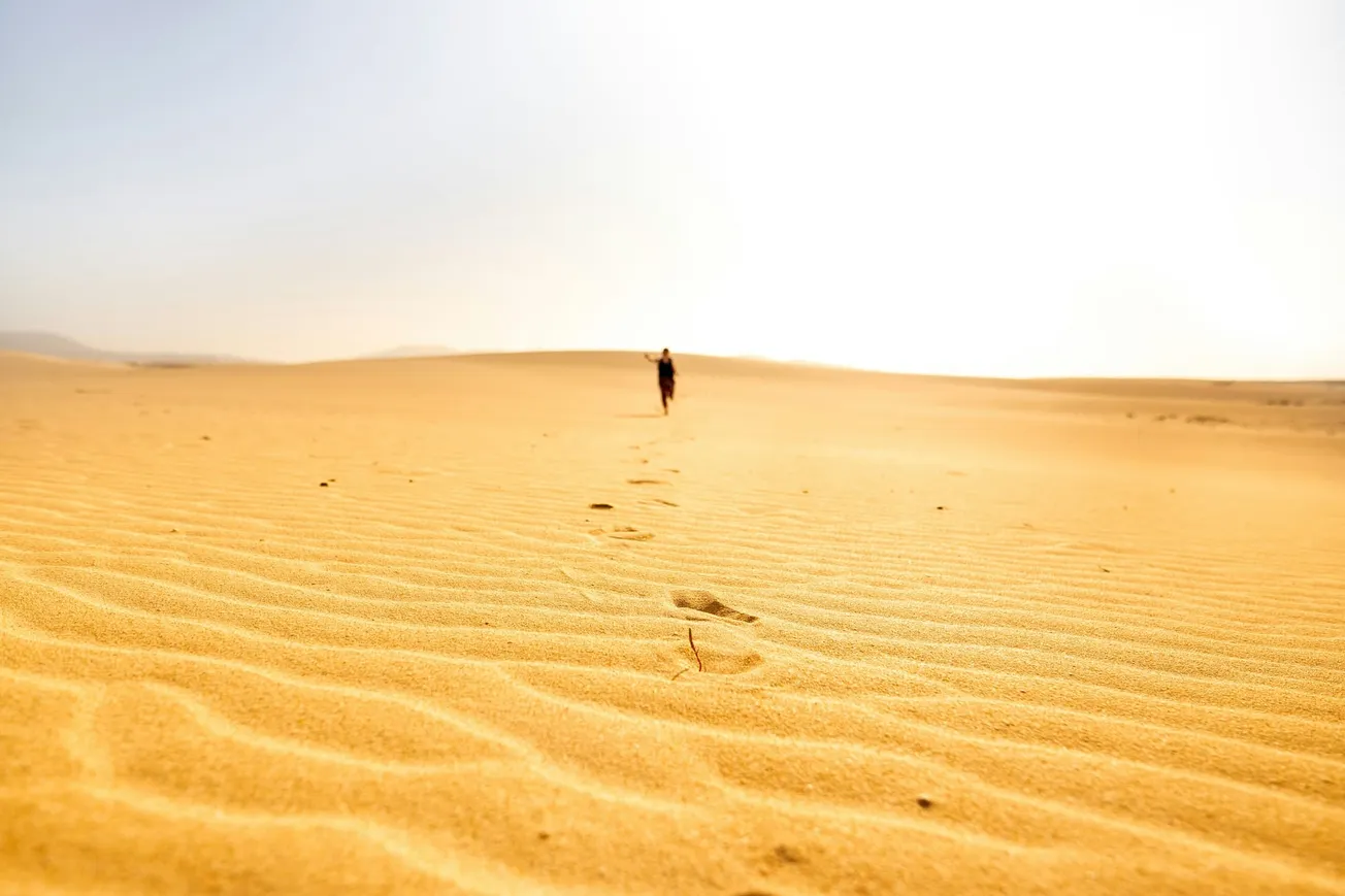 A solitary figure walks across vast, golden sand dunes under a bright, clear sky. Footprints trail behind, evoking a sense of solitude and heat.