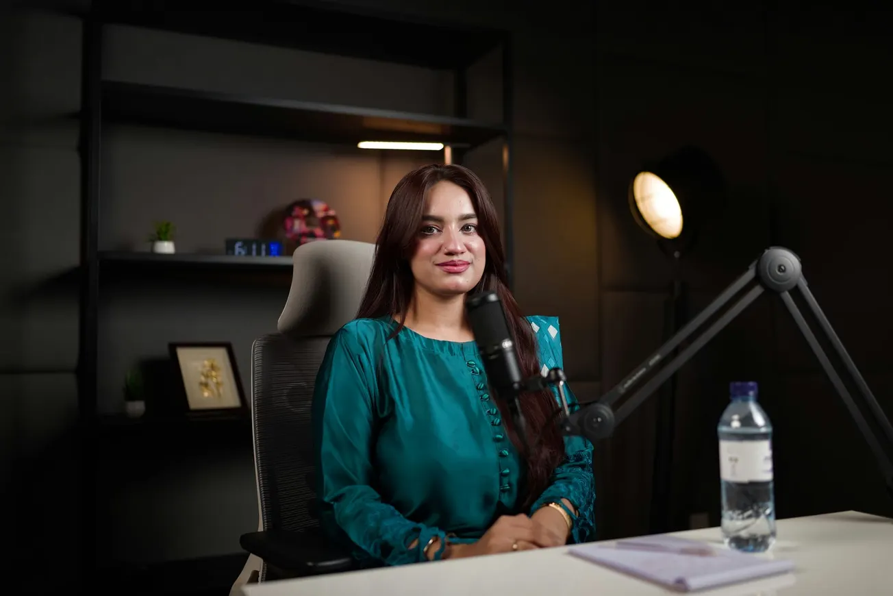 A woman in a teal blouse sits at a desk with a microphone, conveying a podcast setting. The background features shelves, framed art, and soft lighting.