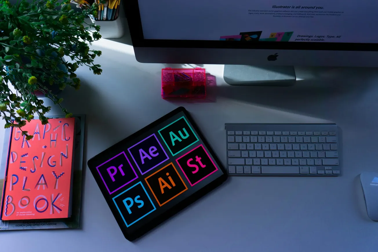 A desktop with a keyboard, monitor, graphic design book, and a tablet displaying colorful Adobe app icons. A plant adds a touch of nature.
