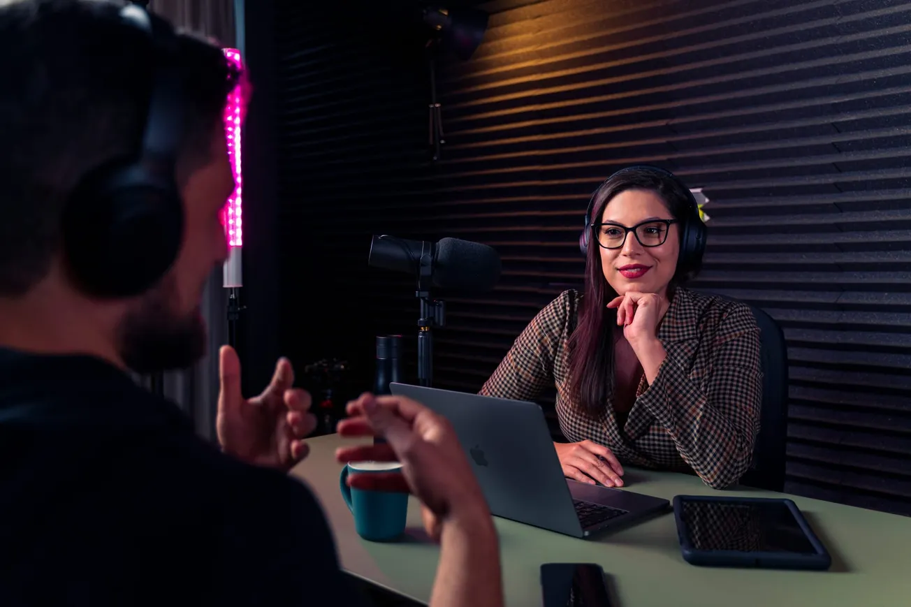Woman in a podcast studio, wearing headphones, sits at a table with a laptop, microphone, and tablet. Engaged, she listens to a man speaking.