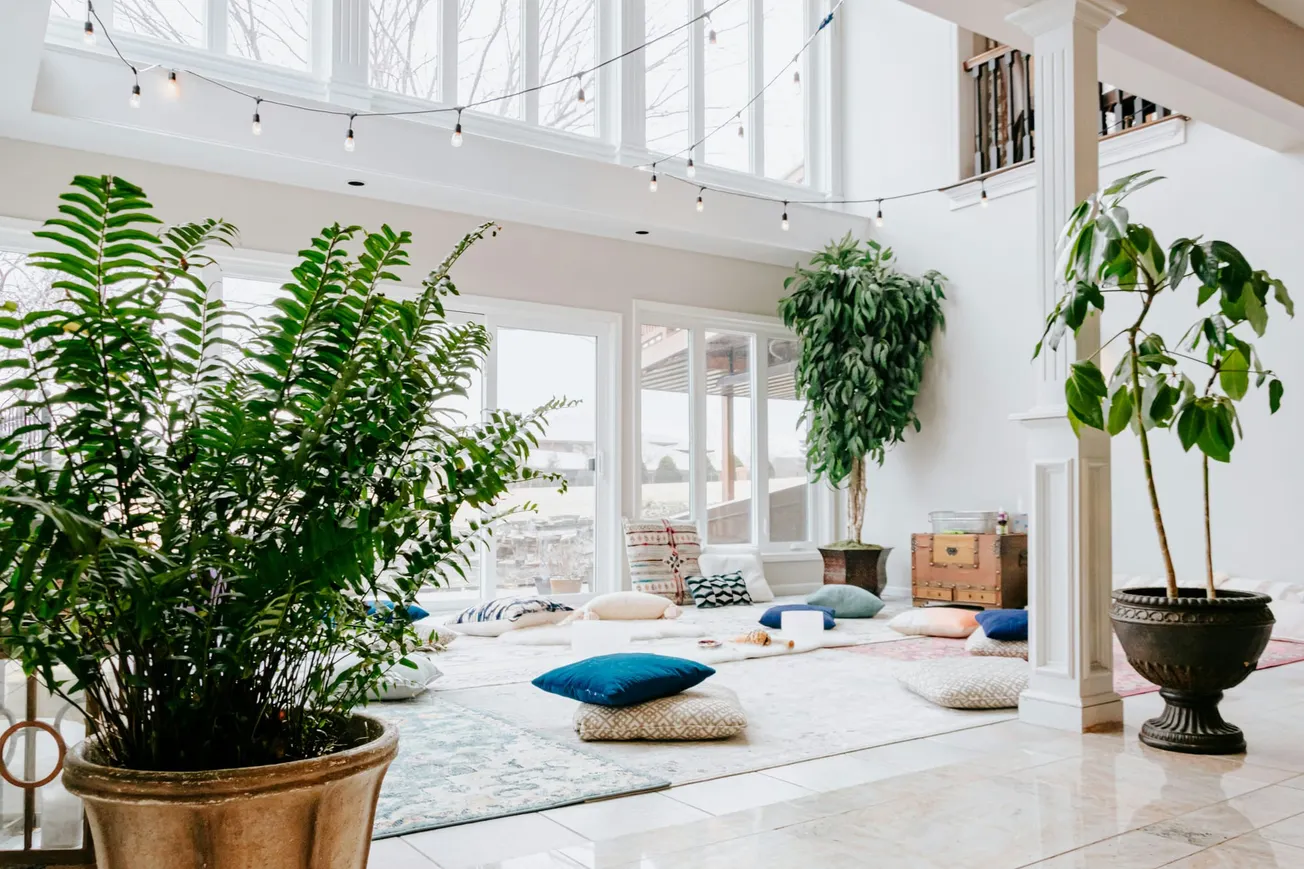 Bright, spacious room with large windows, potted plants, and string lights. Cushions and rugs create a cozy, inviting atmosphere for relaxation.