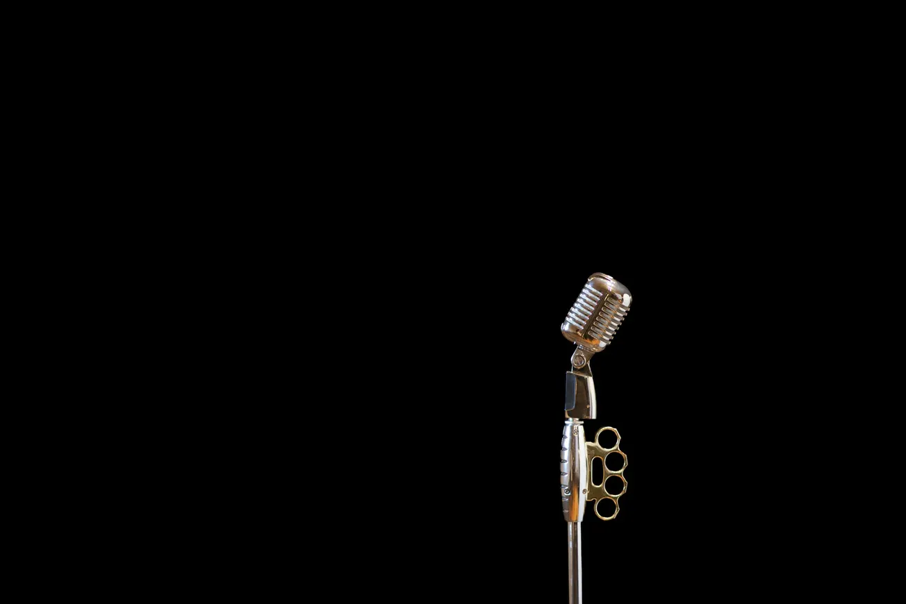 Vintage-style microphone on a stand with brass knuckles attachment, set against a stark black background. Conveys a bold, edgy vibe.