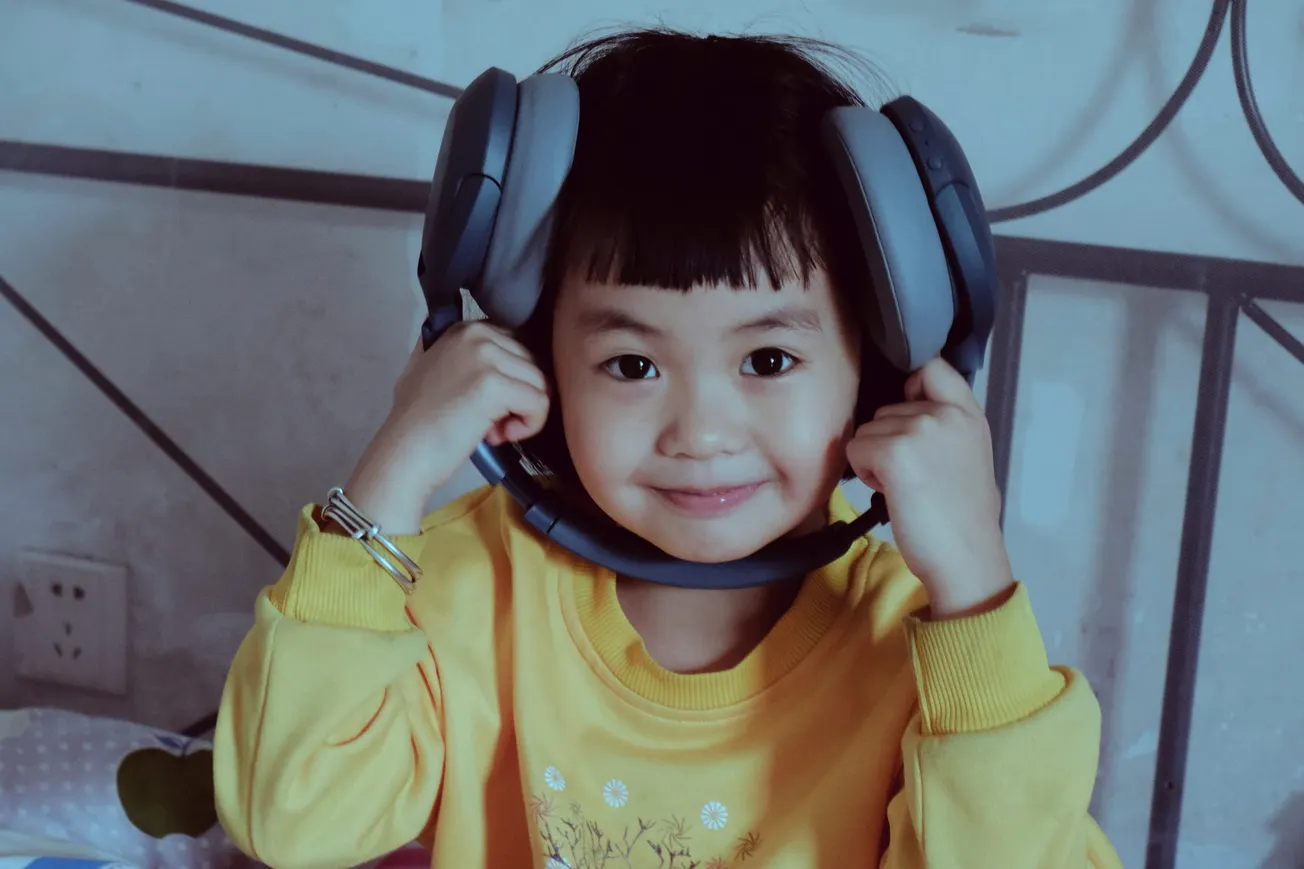 Young child in a yellow shirt holds large headphones around their neck, smiling playfully. The setting is cozy, with warm tones and a relaxed vibe.