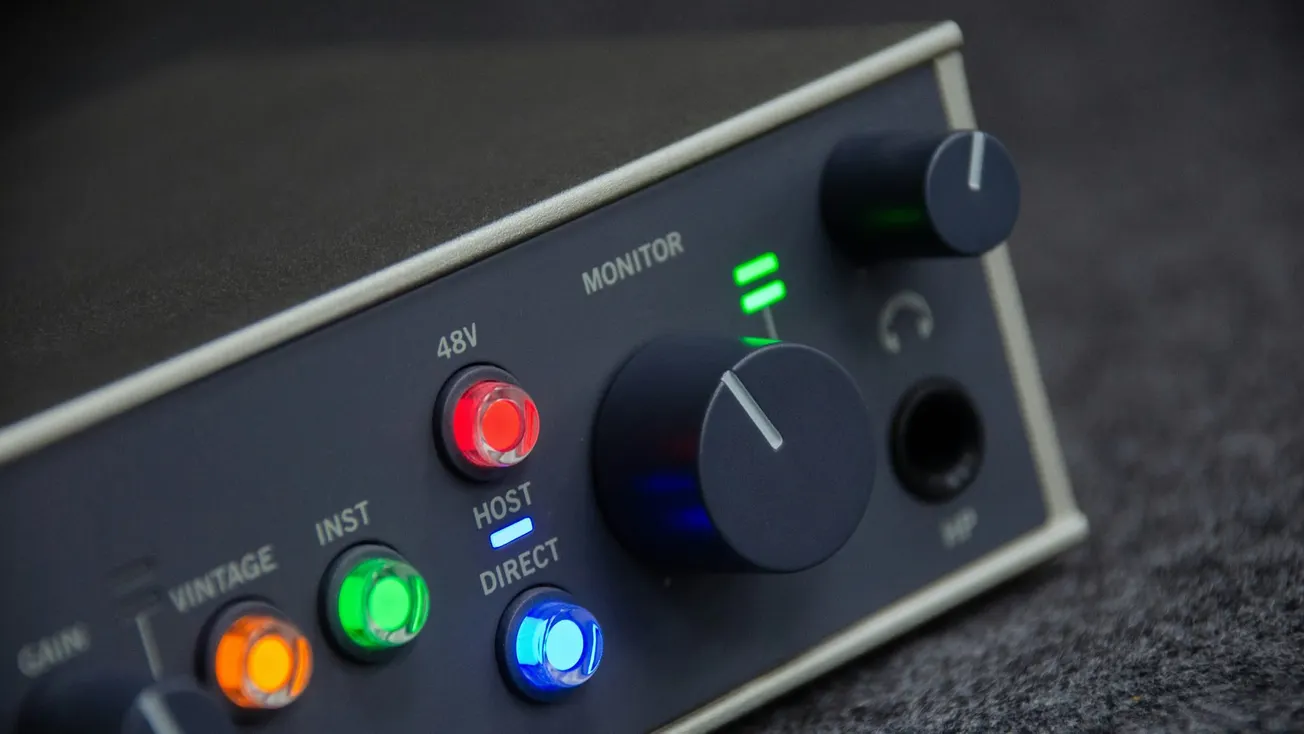 Close-up of an audio interface with colorful LED lights glowing in red, green, and blue. A large knob labeled "Monitor" is prominently featured.