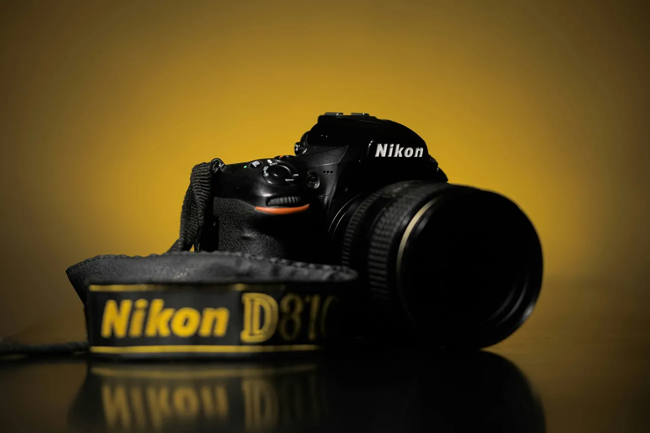 A Nikon D810 camera with a lens sits on a reflective surface against a warm yellow gradient background. The Nikon strap is prominently displayed in front.