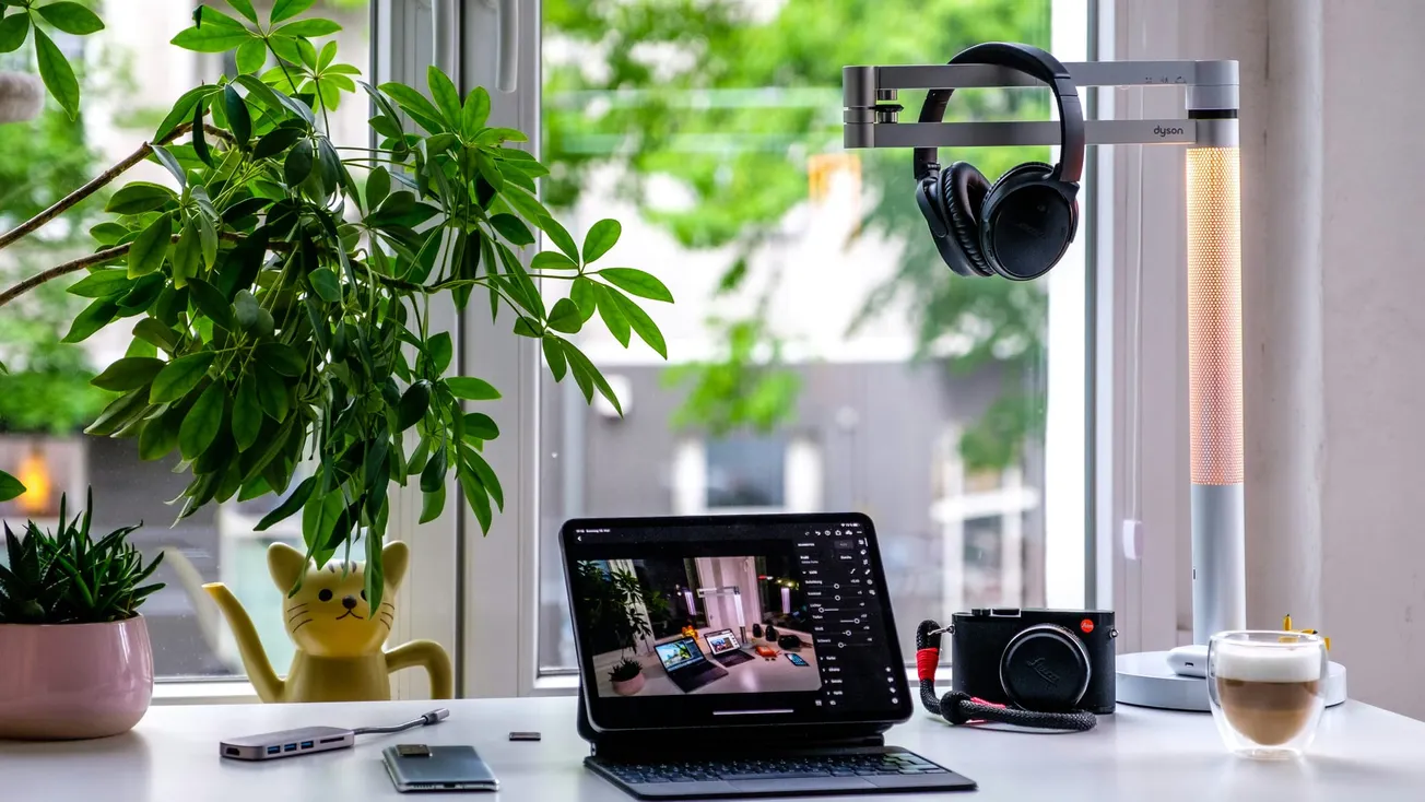 A bright desk by a window features a tablet with a keyboard, a camera, a latte, and a plant. Headphones hang on a sleek lamp, creating a cozy workspace.