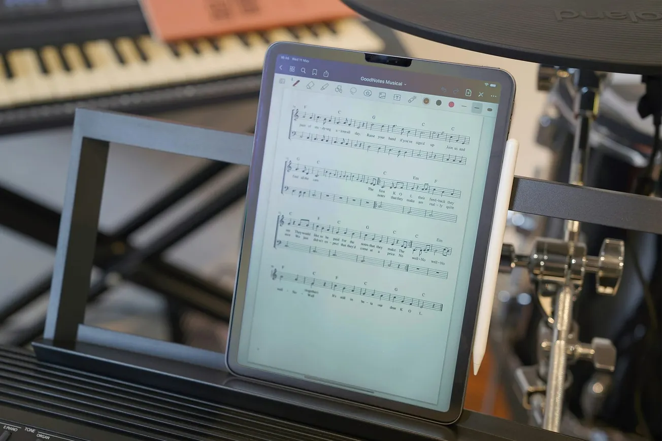 A tablet displaying sheet music is on a stand in a music studio. A drum set and keyboard are blurred in the background, suggesting a creative atmosphere.