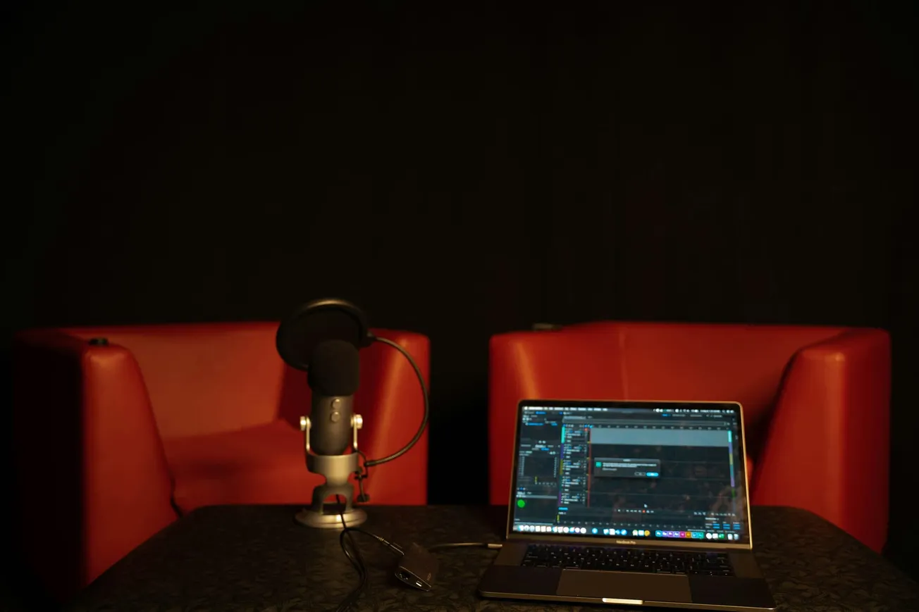 A podcast setup with a microphone on a stand, a laptop displaying audio editing software, and two red armchairs in a dimly lit room.