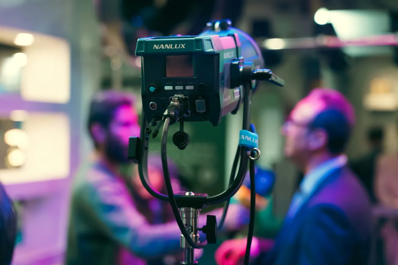 A photography studio with a Nanlux lighting rig in focus. In the blurred background, two people engage in conversation, creating a professional yet casual atmosphere.