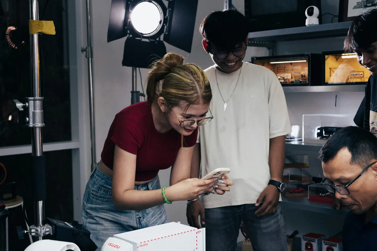 A group of people in a dimly lit room smile and look at a phone. A bright studio light and shelves with equipment are in the background, creating a lively atmosphere.