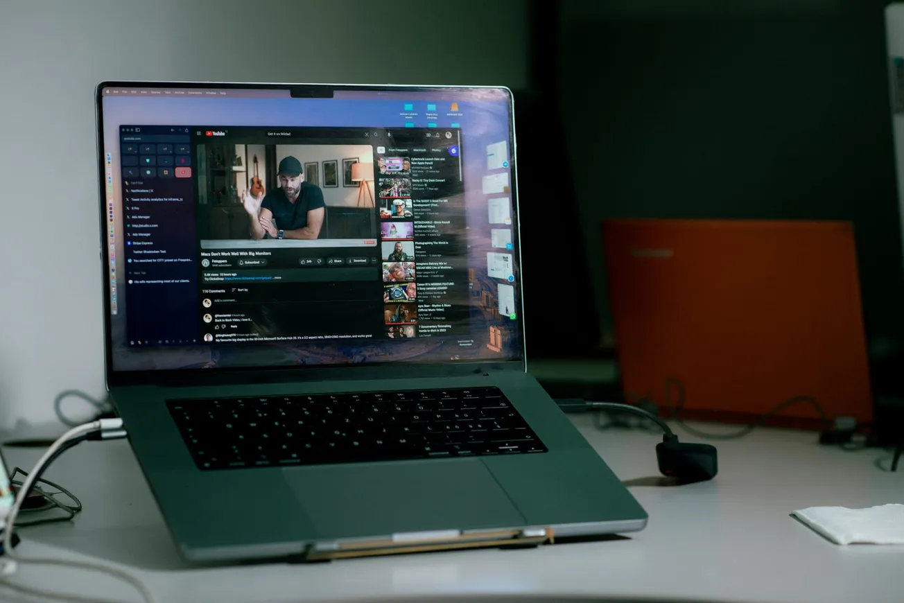 A laptop on a desk displays a YouTube video in a dimly lit room. The screen shows a man sitting, with video suggestions on the right sidebar.