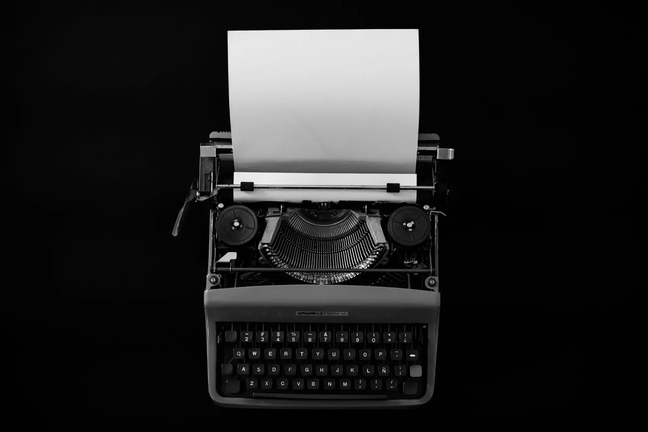 Black and white image of a vintage typewriter with a blank sheet of paper, centered against a dark background, evoking a sense of nostalgia and creativity.