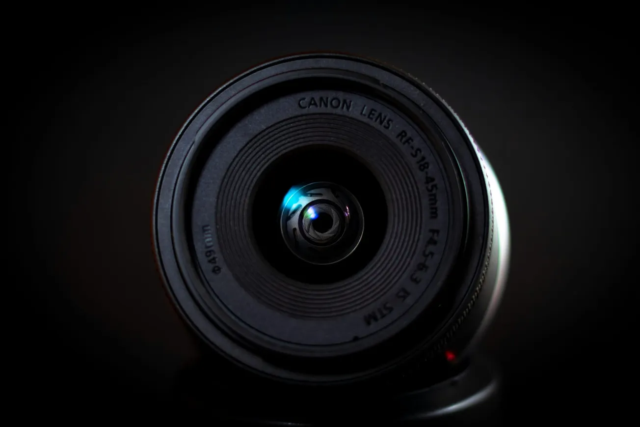 Close-up of a Canon camera lens with visible reflections, showing intricate details of the glass and rings. The background is dimly lit, emphasizing the lens.