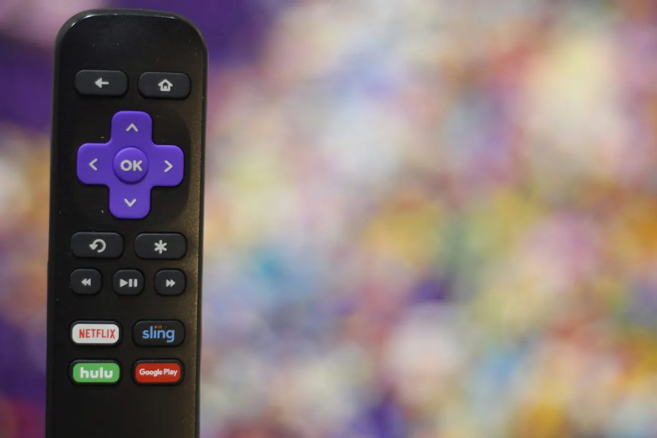 Close-up of a remote control featuring a purple navigation pad and app buttons for Netflix, Sling, Hulu, and Google Play against a colorful blurred background.