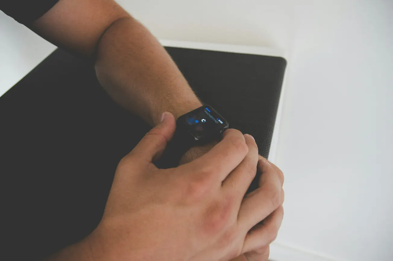 A person is adjusting a smartwatch on their wrist. The watch screen displays blue icons. The setting is minimal, suggesting a modern, tech-savvy vibe.