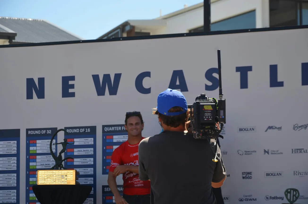 Man in a red shirt smiles for a camera in front of a "NEWCASTLE" banner with a trophy on a pedestal. The scene is outdoors and captures a casual, competitive atmosphere.