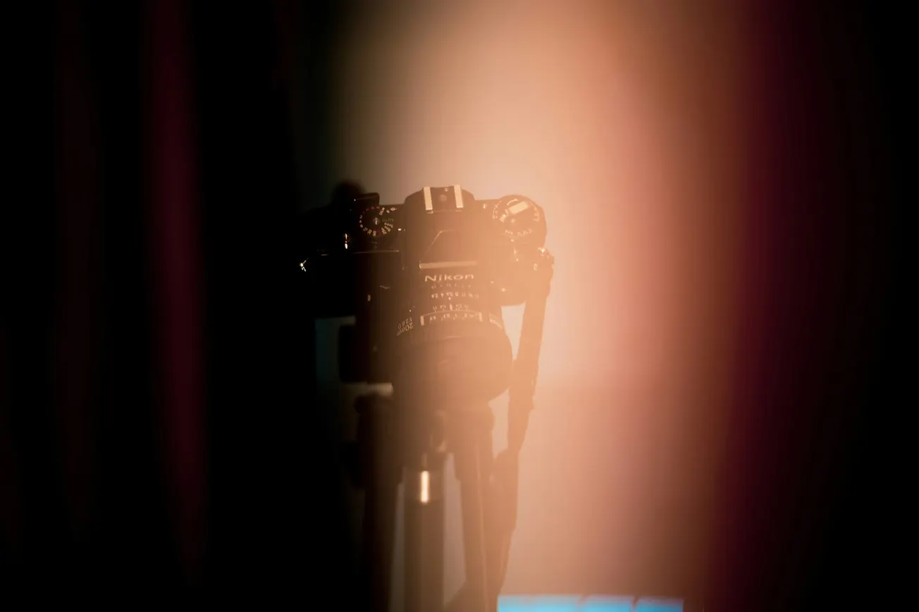 A DSLR camera on a tripod is partially obscured by soft, warm lighting, creating a mysterious and artistic atmosphere with a focus on the lens.
