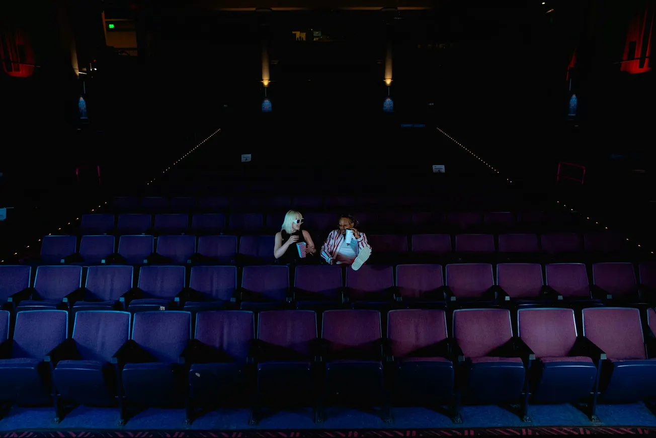 Two people sit in an almost empty, dimly-lit theater, holding popcorn and smiling. Their conversation creates a cozy, intimate atmosphere.