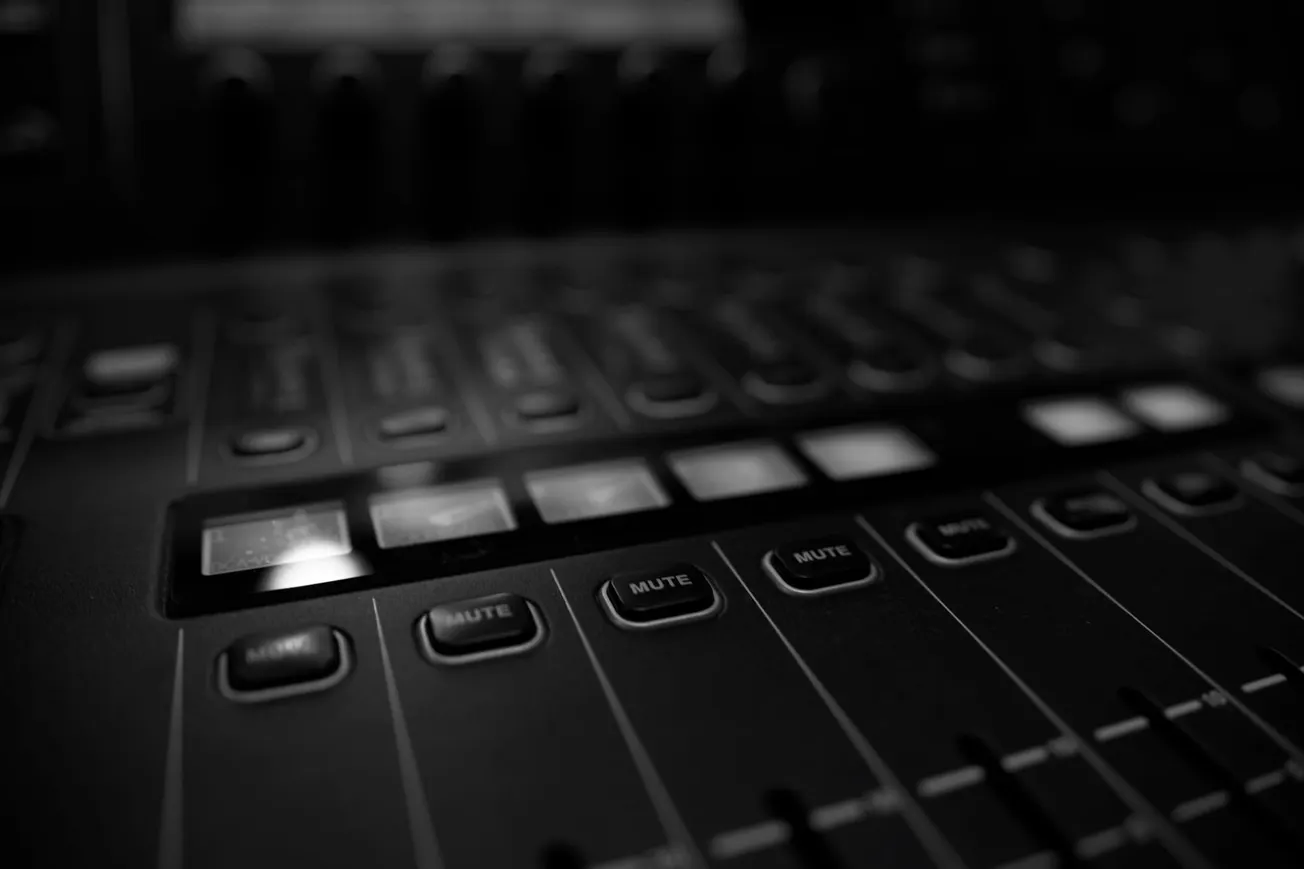 Close-up of a dimly lit audio mixing console with illuminated square buttons and "Mute" switches, conveying a focused and technical atmosphere.