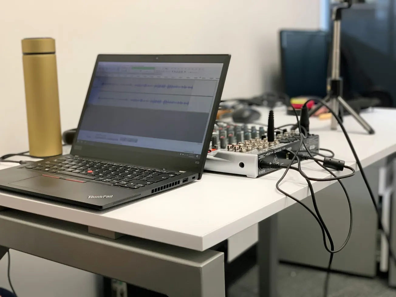 A laptop showing audio software next to a mixer with cables on a desk. A tripod and a gold flask are in the background, suggesting a podcast setup.