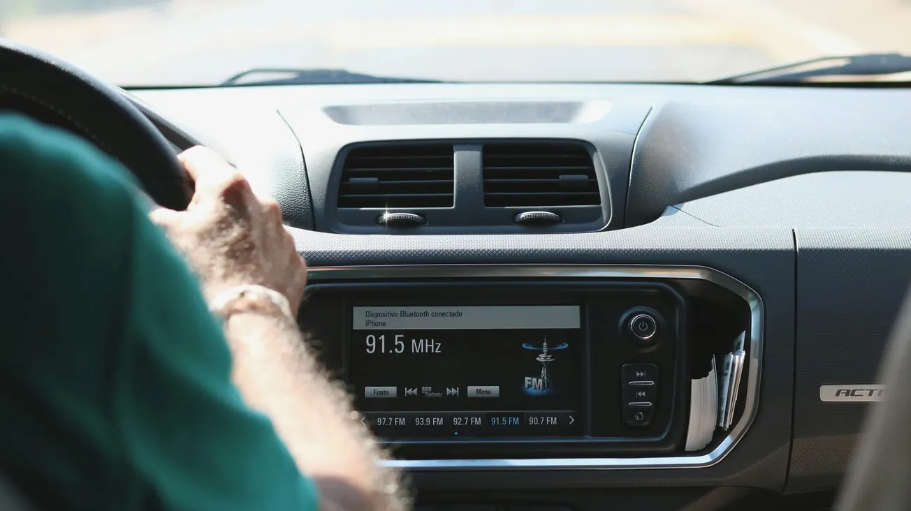 Close-up of a car dashboard with a hand on the steering wheel. The radio display shows a frequency of 91.5 MHz. Daylight, calm drive.