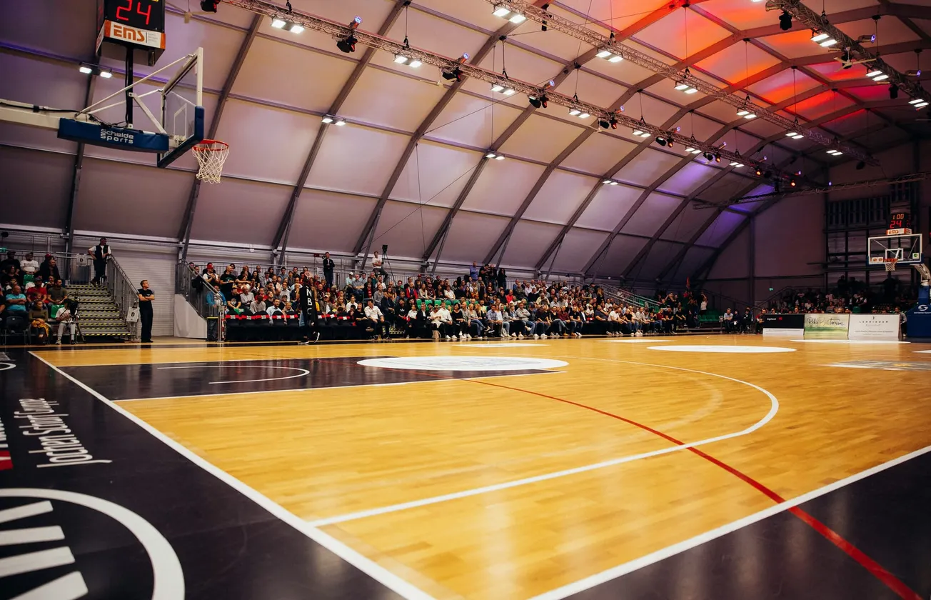 Spacious indoor basketball stadium with wooden court, high ceiling, and full audience. Bright lights and colorful tones create an energetic atmosphere.